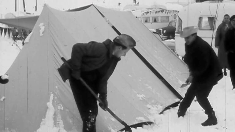 Zwei männlich gelesene Menschen schippen Schnee von einem Zelt