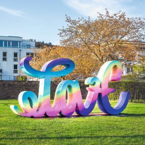 Die farbig lackierte Stahlskulptur "Love Hate" von Mia Florentine Weiss formt von einer Blickrichtung das Wort "Love". Bewegt sich der Standpunkt der Betrachtung auf die andere Seite der Skulptur formt diese das Wort "Hate".