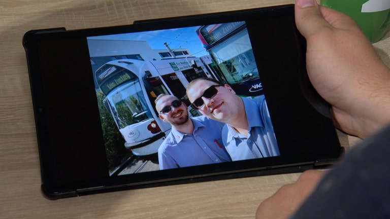 Die Ehemänner und Straßenbahnfahrer Daniel und Marco stehen vor einer Tram und lächeln in die Kamera