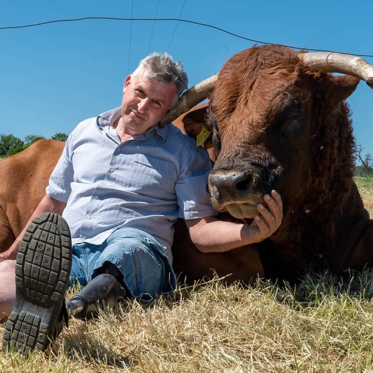 Mann mit Beinprothese einen Bullen streichelnd auf dem Feld sitzend. (Foto: SWR)