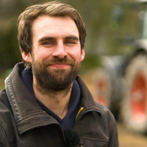 Junger Mann mit langem Bart, brauner Jacke vor einem Traktor (Foto: SWR)