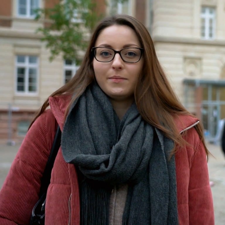 Eine junge Frau mit braunen, glatten Haaren und Brille schaut neutral in die Kamera