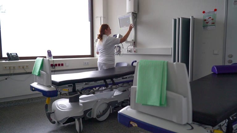 Junge Frau mit roten Haaren und weißer Arbeitskleidung in einem Krankenhausbehandlungsraum. Sie stellt einen Monitor ein.