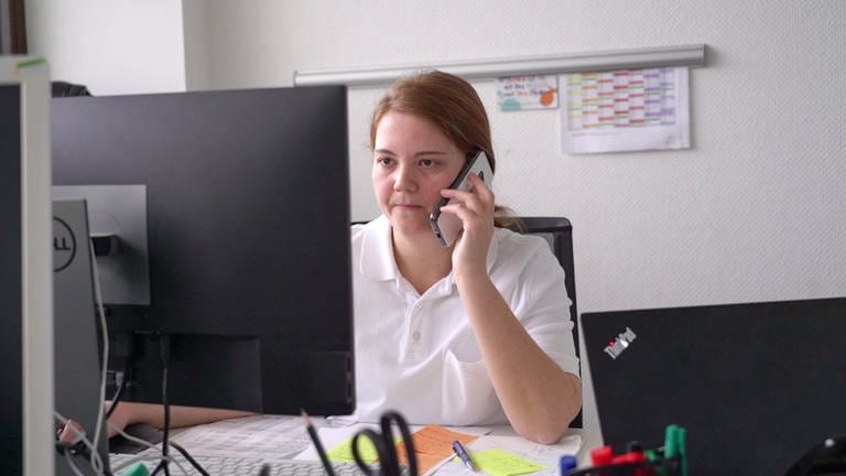 Junge Frau mit roten Haaren und weißem Polo-Shirt sitzt an einem Computer und telefoniert mit einem Handy. 
