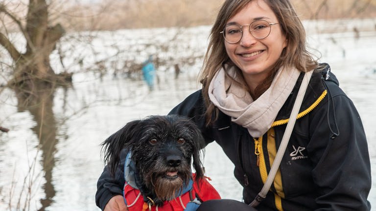 Hund und Frau mit Brille lächeln vor Fluss.