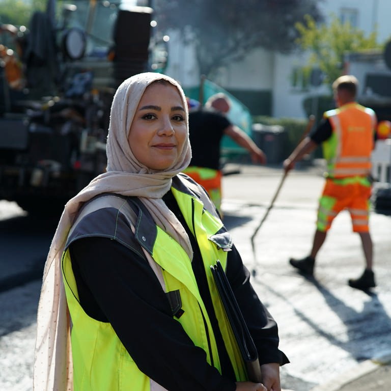 Bauingenieurin Hawra lächelt in die Kamera und steht auf einer Baustelle. Im Hintergrund sind Straßenbauarbeiter zu sehen. (Foto: SWR)