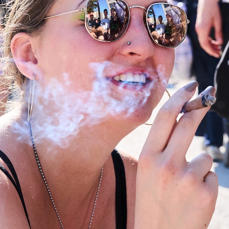 Junge Frau mit Sonnenbrille raucht einen Joint. Ab 1. April ist der Konsum und Besitz von Cannabis in bestimmten Mengen erlaubt. | Was bringt die Teil-Legalisierung von Cannabis?