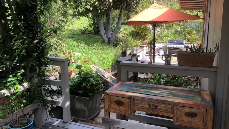 Kleiner Holz-Schreibtisch auf der Terrasse, Blick in den Garten mit alten Bäumen, Blumen und auf einen orange-farbenen Sonnernschirm (Foto: SWR, privat)