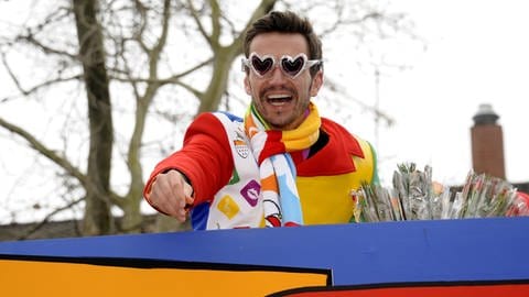 Florian Silbereisen trägt eine bunte Uniform und eine herzförmige Brille. Er Ist auf einem Karnevalswagen und jubelt der Menge zu.