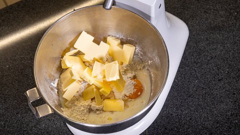 Die Zutaten für einen "All-in-Hefeteig" befinden sich in eine Rührschüssel. Darunter Hefe, Butter, Mehl, Eier und Salz.