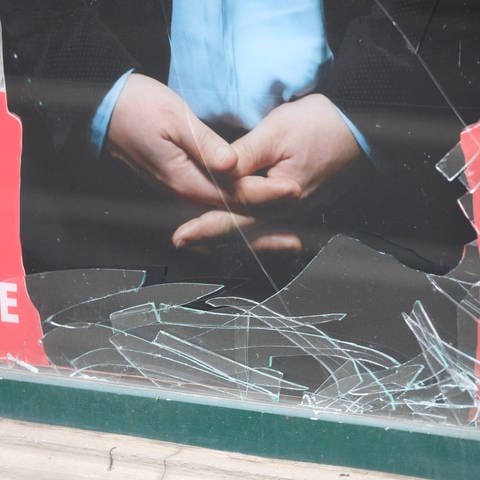 Ein Plakat hängt in einem eingeschlagenen Schaufenster. Überall sind Glasscherben zu sehen.