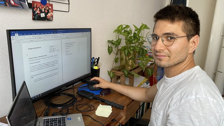 Karim Baghlani am Schreibtisch vor Computer mit Maus in der Hand und Pflanze im Hintergrund