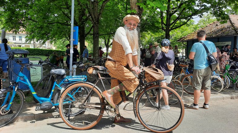 Ein Mann mit langem weißem Bart in karierter Kleidung auf einem Retro-Bike lacht in die Kamera. (Foto: SWR)