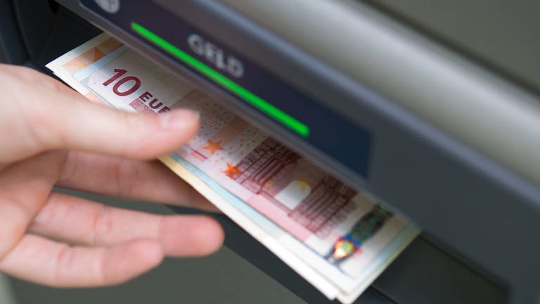 Eine Hand greift nach Geldscheinen, die aus einem Geldautomaten ausgeworfen wurden. 
