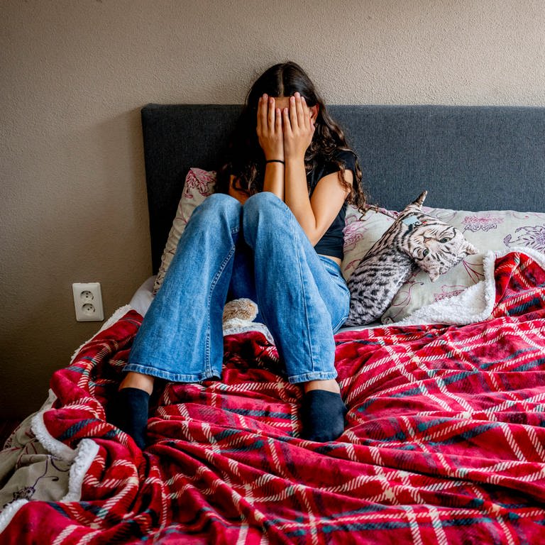 Jugendliches Mädchen mit Depression sitzt auf ihrem Bett und hält sich die Hände vors Gesicht