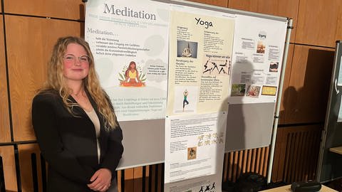 Eine junge Frau steht vor verschiedenen Plakaten. Eine Schülerin der Julius-Wegeler-Schule stellt verschiedene Entspannungsübungen vor, die sie in der Schule erlernt hat. (Foto: SWR)
