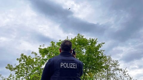 Ein Pressesprecher der Polizei in Mainz steht mit einem Funkgerät am Rande der Gefahrenzone. Am Himmel kreist der Polizeihubscharuber. 