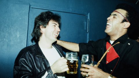 Frank Farian und Bobby Farrell, eigentlich Roberto Alfonso Farrell, von Boney M. in den 1980er Jahren in Köln. Beide haben Glasbierkrüge in der Hand, links steht Farian, rechts Farrell. Farian trägt eine schwarze Lederjacke über einem weißen Shirt, Farrell eine schwarz-rote doppelseitig geknöpfte Jacke, er hat auch ein rot-goldenes schmales Stirnband.