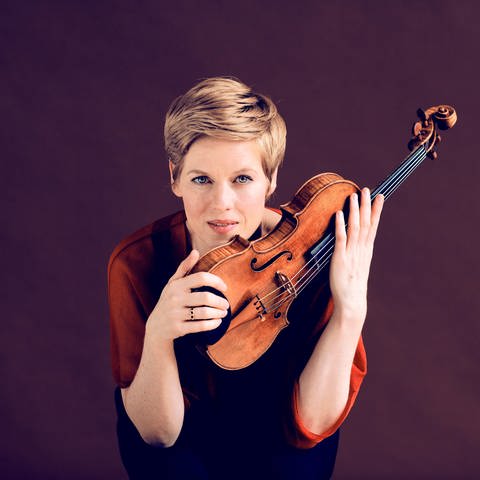 Isabelle Faust (kurzes, blondes Haar) mit Geige vor violletem Hintergrund