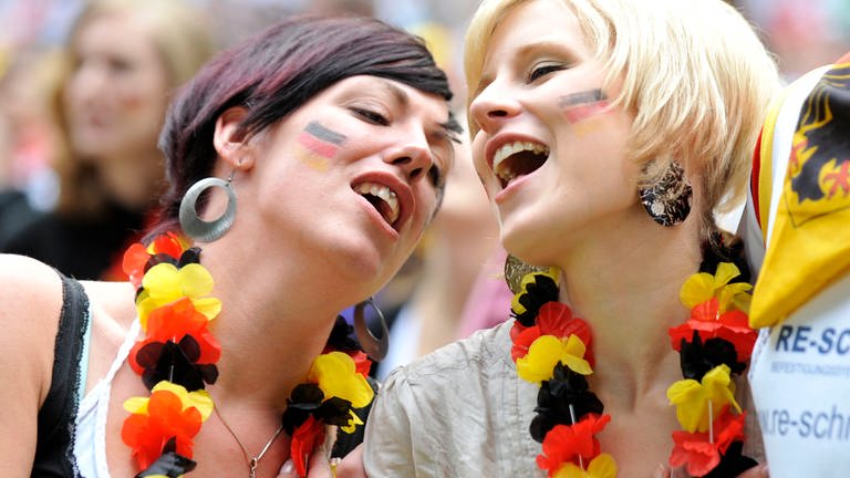 Zwei junge Frauen mit Blumenkette in Deutschland-Farben und der Hand auf dem Herzi singen beim Fußballspiel in Düsseldorf 2010 die Nationalhymne