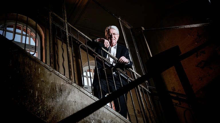 Frank Elstner steht in einem Treppenhaus, lehnt am Geländer und blickt nach unten. 
