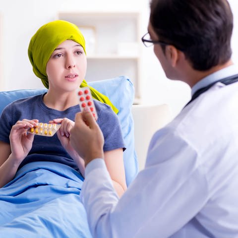 Frau mit Glatze unter ihrem gelben Kopftuch redet mit Arzt über ihre Chemotherapie: Eine Chemotherapie hat oft Haarausfall als Nebenwirkung