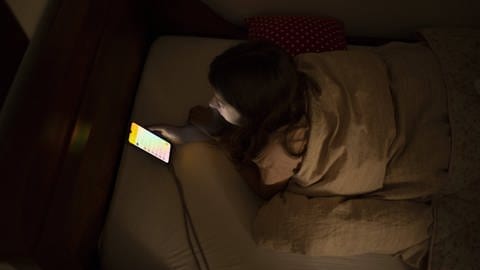 Frau liegt mit Handy im Bett, tags: innere Uhr, Bildschirmnutzung, Schlafenzeit