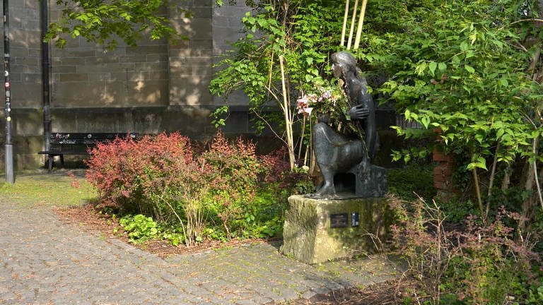Man sieht eine Skulptur. Sie steht am Rand eines kleinen Weges in Tübingen. Man nennt sie: die "Sitzende". SIe hat einen Strauß Blumen im Arm. Die Blumen sind schon etwas verwelkt. (Foto: SWR)