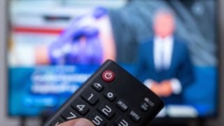 Eine Hand tippt auf eine TV-Fernbedienung, im Hintergrund unscharf der dazugehörige Fernseher, auf dem die tagesschau läuft.