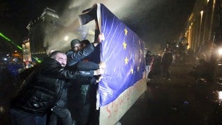 Menschen in Tiflis halten einen Teil eines Zauns mit einer EU-Flagge, während die Polizei einen Wasserwerfer einsetzt.