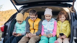 Kinder mit Mobiltelefonen sitzen in der offenen Kofferraumtür eines PKWs.