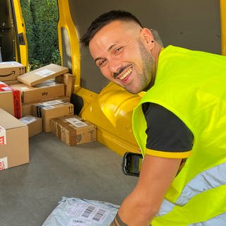 DHL-Postbote Vincenzo lädt Pakete aus dem Postauto aus. Vincenzo strahlt und hat gute Laune.