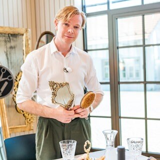 Friseur Dennis ist in seinem Salon und hat eine Schere, Bürste und einen Spiegel in der Hand und liebt seinen Job. Er schaut direkt in die Kamera.