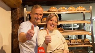 Hausarzt Jörn und seine Tochter Nora stehen lachend vor der Auslage, in der frische Brote liegen, in ihrer Konstanzer Bäckerei. Sie machen beide einen Daumen nach oben. 