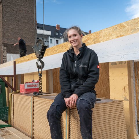 Eine junge, blonde Frau sitzt lächelnd in Arbeitskleidung auf einem Stapel OSB-Platten auf einer Baustelle.