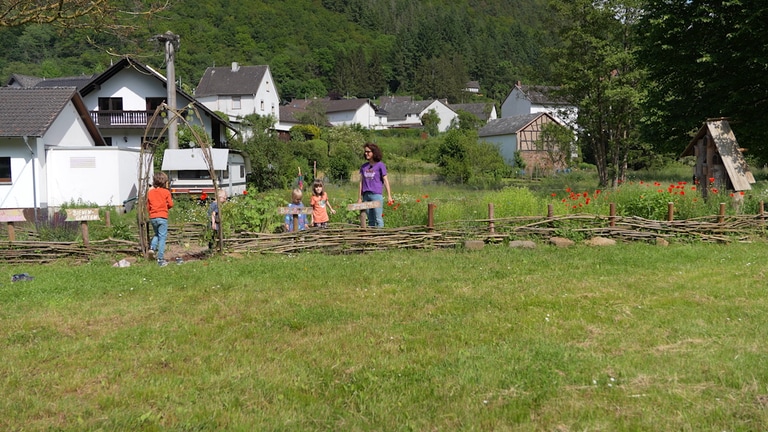 Bienengarten mit Frau und Kindern im Vordergrund. Im Hintergrund das Dorf.