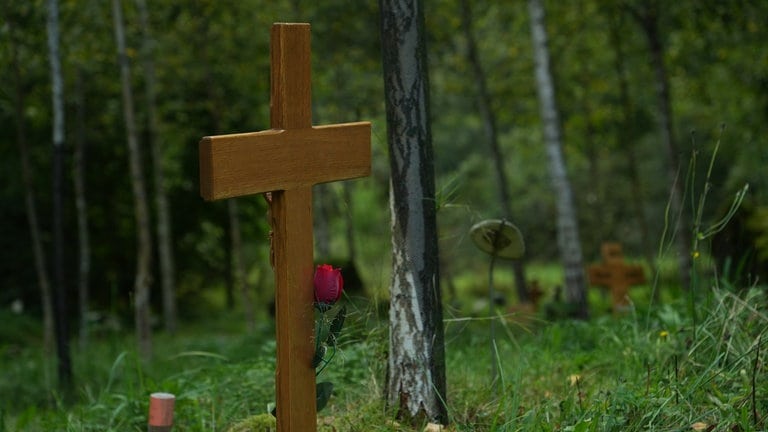 Holzkreuz mit einer Rose vor einem Baumstamm und Wiese.