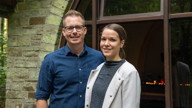 Ein blonder Mann mit Brille und dunkelblauem Hemd und eine Frau mit braunen Haaren und weißem Mantel lächeln in Kamera.