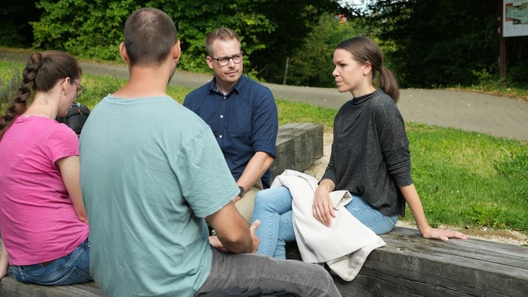 Zwei Männer und zwei Frauen sitzen sich auf Holzbänken gegenüber.