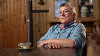 Eine mittelalte Frau mit blauem Hemd, Halstuch und kurzen grauen Haaren blickt in Richtung Kamera. Sie sitzt in einem Bauernhaus und erzählt von ihrer Zeit im Kinderheim.