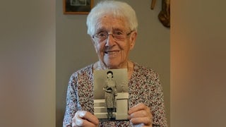 Alte Frau mit weißen Haaren und Brille lächelt in Kamera und hält ein schwarz-weiß Foto in den Händen.