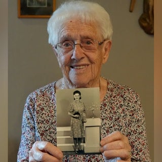 Alte Frau mit weißen Haaren und Brille lächelt in Kamera und hält ein schwarz-weiß Foto in den Händen.