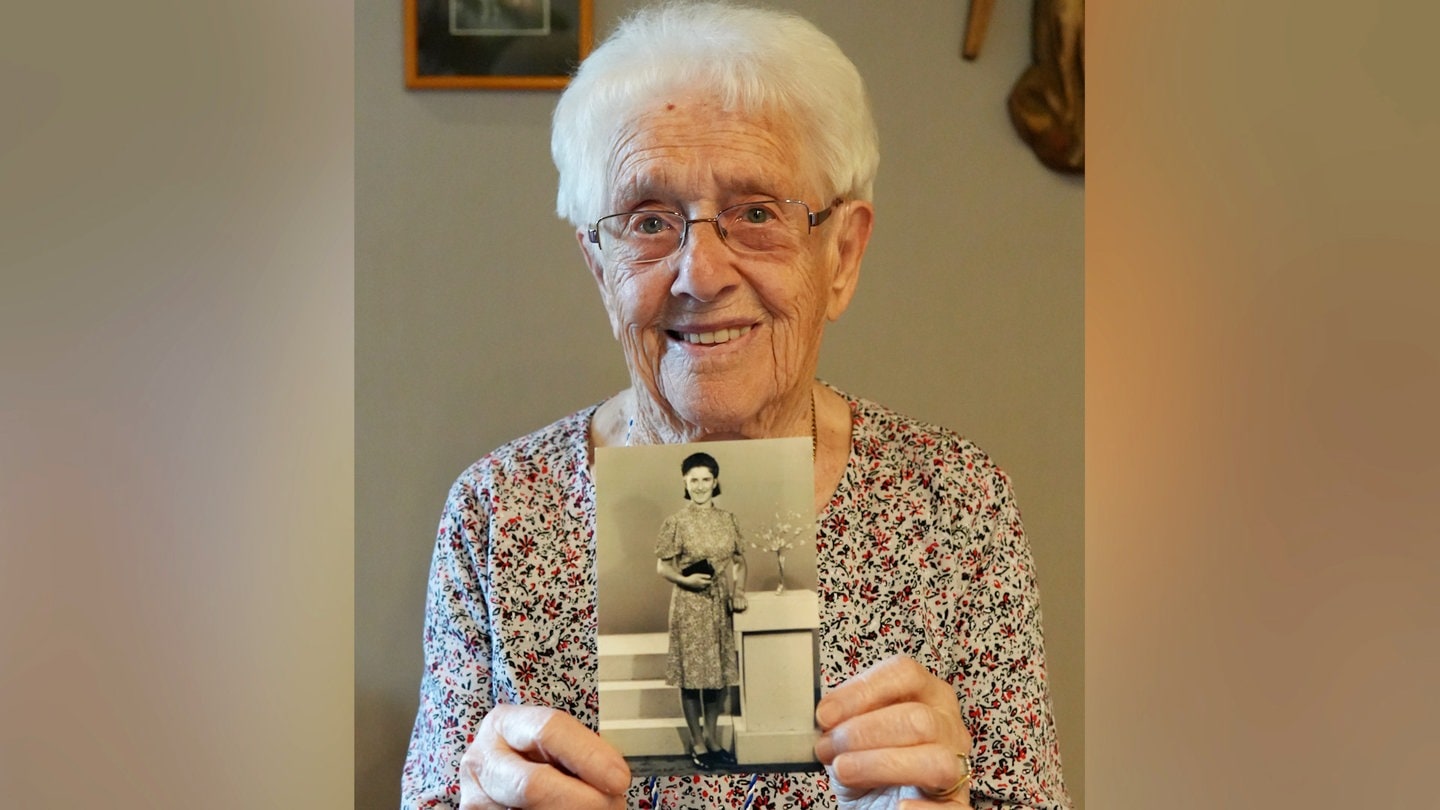 Alte Frau mit weißen Haaren und Brille lächelt in Kamera und hält ein schwarzweiß Foto in den Händen.