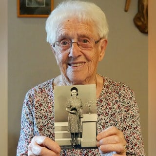 Alte Frau mit weißen Haaren und Brille lächelt in Kamera und hält ein schwarzweiß Foto in den Händen.