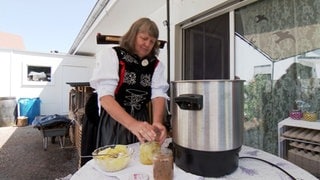 Eine Frau in Tracht steht draußen an einem Tisch mit großem Topf und schließt ein Schraubverglas mit Gemüse 