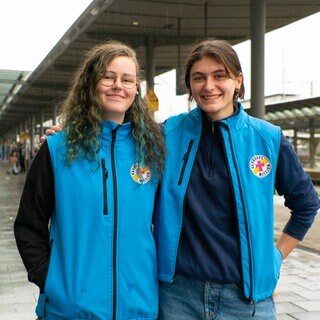 Sinja und Ellen machen ihr Freiwilliges Soziales Jahr bei der Bahnhofsmission in Freiburg. Sie tragen die Arbeitskleidung der Bahnhofsmission und lachen in die Kamera. 