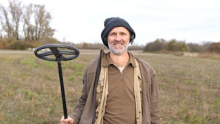 Gerhard ist Hobby Archäologe und hält einen Metalldetektor in der Hand und lächelt in die Kamera.