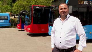 Hazim ist Busfahrer in Biberach. Er trägt ein weißes Hemd. Im Hintergrund sind drei Busse zu sehen: Einer ist blau die anderen beiden rot. Hazim lacht in die Kamera.