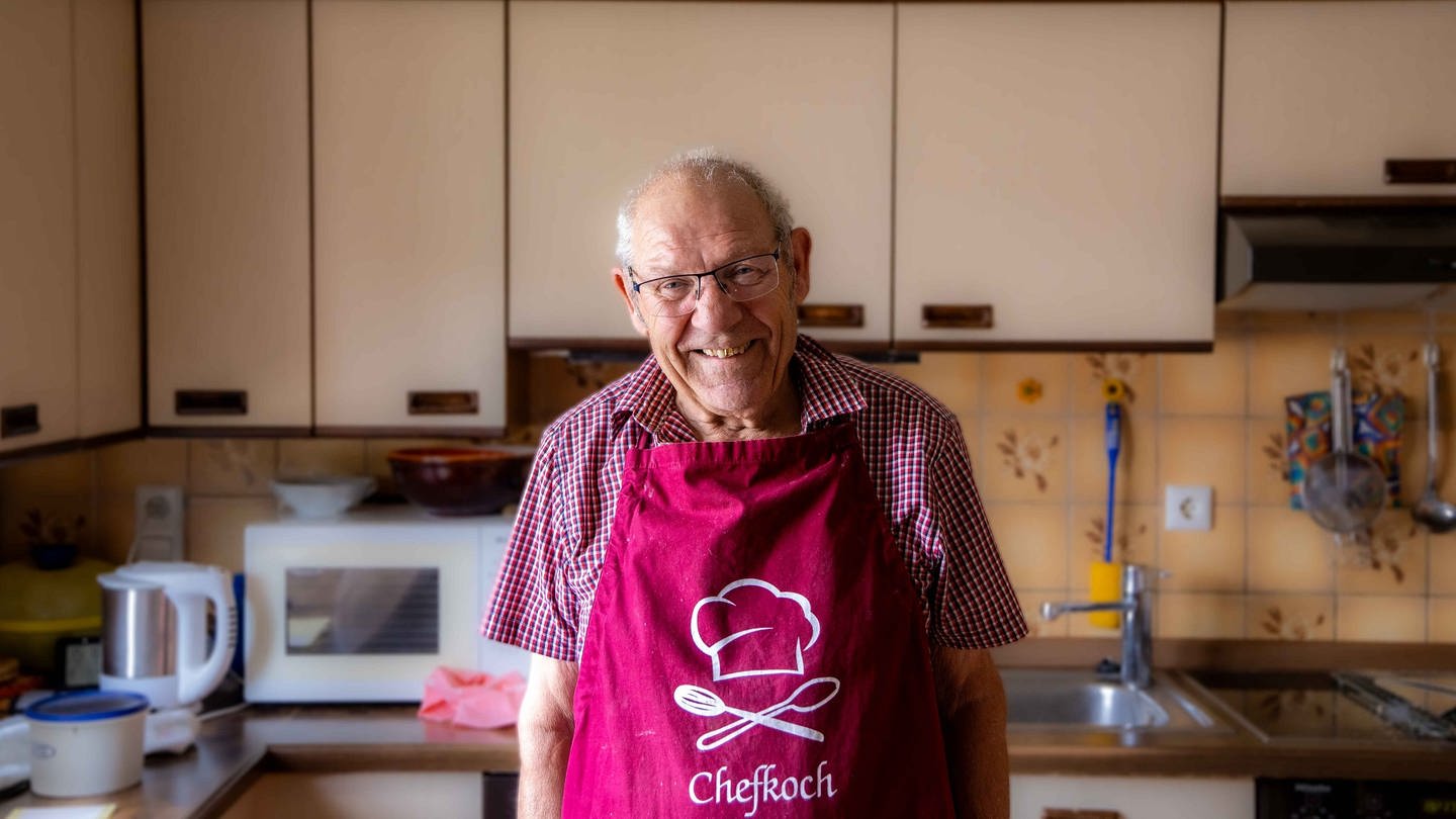 Ältere Herr mit Brille und Kochschürze steht in seiner Küche und lächelt in Kamera.