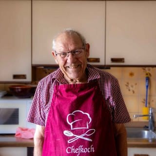 Ältere Herr mit Brille und Kochschürze steht in seiner Küche und lächelt in Kamera.